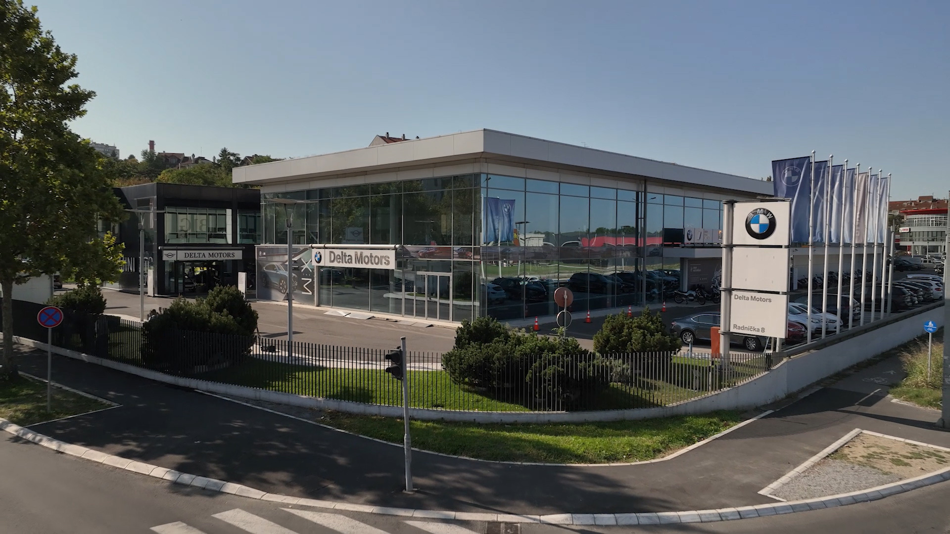 Auto salon BMW & MINI — širok kadar salona sa staklenom fasadom, zelenilom i BMW zastavama