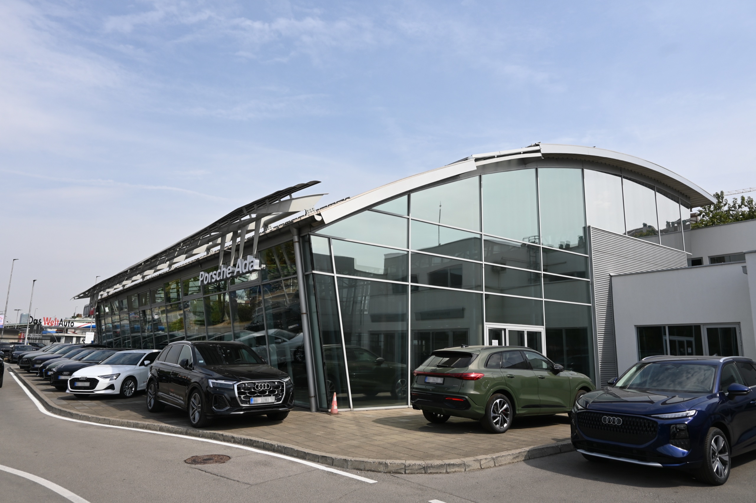 Auto salon Porsche Ada — staklena fasada pod zakrivljenim krovom i nadstrešnica od lamela, automobili ispred